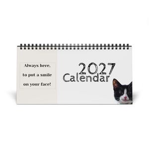May include: A horizontal 2027 desk calendar with a black spiral binding. The cover features a black and white cat face on the right and the text Always here, to put a smile on your face! printed in a box on the left side of the white background.