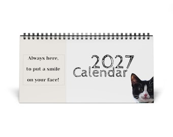 Puede incluir: Un calendario de escritorio horizontal 2027 con encuadernación en espiral negra. La portada muestra la cara de un gato blanco y negro a la derecha y el texto Always here, to put a smile on your face! impreso en un recuadro sobre el fondo blanco.