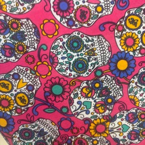 Puede incluir: Una tela rosa vibrante con un patrón repetido de diseños de calaveras de azúcar. Cada calavera está intrincadamente decorada con detalles florales y ornamentales coloridos en tonos de azul, amarillo, rosa y verde. El diseño es un textil festivo y llamativo.