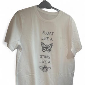 May include: White t-shirt featuring the text "FLOAT LIKE A" above a butterfly illustration, and "STING LIKE A" above a bee graphic. The shirt is suspended on a black hanger.