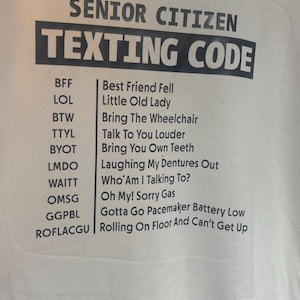 May include: White t-shirt featuring a black graphic with the text "SENIOR CITIZEN TEXTING CODE." The graphic lists texting acronyms and their meanings, including "LOL" for "Little Old Lady" and "ROFLACGU" for "Rolling On Floor And Can't Get Up."