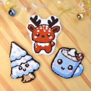 以下が含まれることがあります： 手作りのクリスマスをテーマにした3つの装飾品。茶色の角と白い斑点のあるトナカイ、青と白のクリスマスツリー、ホイップクリームが入った青いマグカップ。すべて黒いアウトラインと可愛い顔をしています。