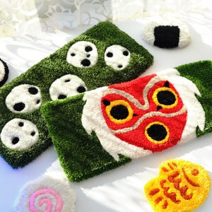 May include: A selection of colourful, plush, handmade items. Includes a green rectangular rug with white and black circular designs, a red and white Pokeball, and a red, white, and yellow mask-like design.