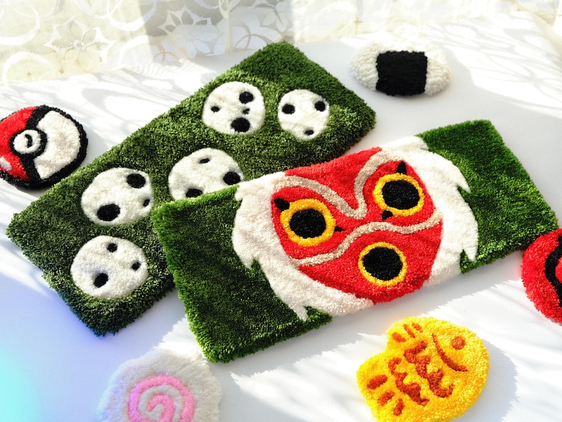 May include: A selection of colourful, plush, handmade items. Includes a green rectangular rug with white and black circular designs, a red and white Pokeball, and a red, white, and yellow mask-like design.