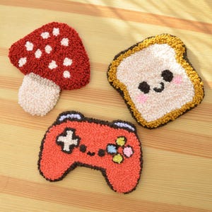 Puede incluir: Tres posavasos hechos a mano, coloridos y texturizados. Uno es una seta roja con lunares blancos, otro es una rebanada de pan con una cara sonriente y el último es un mando de videojuegos rojo.