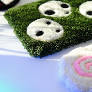 May include: A green, textured rug with white and black panda face designs. A white rug with a pink and white spiral design is also visible. The rugs are made of a soft, plush material.