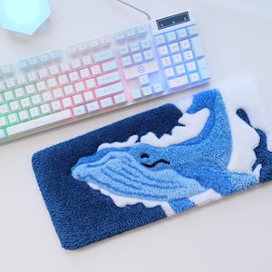 Peut inclure: Un repose-poignet de clavier tufté rectangulaire présentant un motif de baleine bleue texturé sur un fond bleu foncé avec des accents de mousse blanche. Le repose-poignet est placé sous un clavier mécanique blanc avec un rétroéclairage arc-en-ciel.