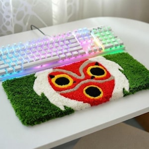 May include: A white mechanical keyboard with multi-coloured LED lights rests on a green and red rug with a character design. The rug has a green border and a red and white face design.
