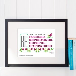 May include: A framed cross-stitch design with the text "BE DON'T BE AFRAID. FOCUSED. DETERMINED. HOPEFUL. EMPOWERED." and a row of pink tulips at the bottom. The text is in a green and gray font. The background is white.