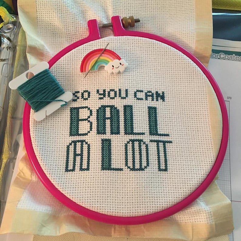 May include: A pink embroidery hoop with white fabric inside. The fabric has a green cross-stitch design that reads "So you can ball a lot". A rainbow and cloud embroidery charm is attached to the fabric.