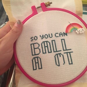 May include: A pink embroidery hoop with white fabric inside. The fabric has a cross-stitch design that reads "so you can ball out". A rainbow-colored pin with a cloud is attached to the fabric.