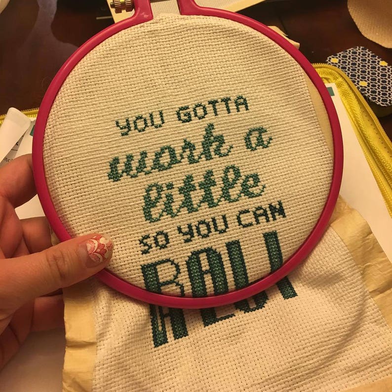 May include: A white embroidery hoop with a green cross-stitch design that reads "You gotta work a little so you can roll".
