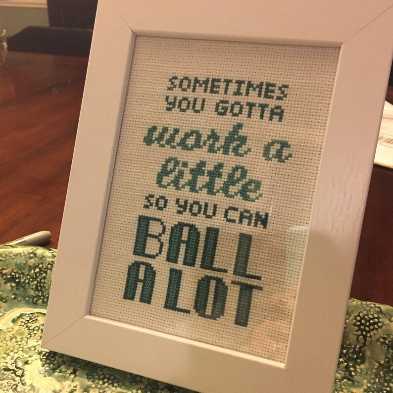 May include: White framed cross-stitch with green text that reads "Sometimes you gotta work a little so you can ball a lot."