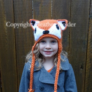May include: A young girl wearing a crocheted orange fox hat with white accents and long orange tassels. The hat has black eyes and a black nose. The girl is wearing a grey coat.