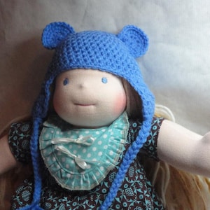 May include: A handmade doll with a blue crocheted bear hat and a blue and white polka dot bib. The doll has long blonde hair and is wearing a brown and blue patterned dress. The doll's face is soft and has rosy cheeks.
