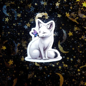 May include: A white fox sticker with a butterfly on its nose. The fox is sitting and winking. The sticker is on a black background with gold and silver stars and crescent moons.
