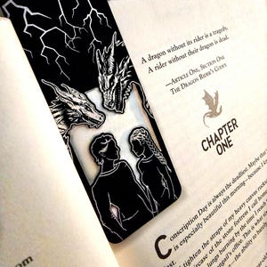 May include: A rectangular black bookmark featuring a white line art illustration of two dragons at the top and two human figures facing each other at the bottom. The top section of the bookmark displays a white lightning bolt pattern against a black background.