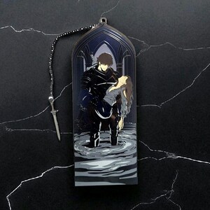 May include: A metal bookmark featuring a dark illustration of a figure in black armor carrying a woman in a flowing gown. The bookmark has a chain and a small sword charm. The background depicts an arched gothic structure.
