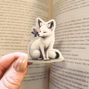 K&ouml;nnte beinhalten: Ein wei&szlig;er Fuchs-Aufkleber mit schwarzspitzem Schwanz und Ohren sowie einem lila Schmetterling auf der Nase. Der Fuchs sitzt vor einem offenen Buch.