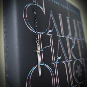 May include: A silver sword charm hangs from a black chain, resting against a dark book cover. The book's title is in large, stylized letters with a blue and pink gradient. The text at the top reads "The #1 New York Times Bestseller."