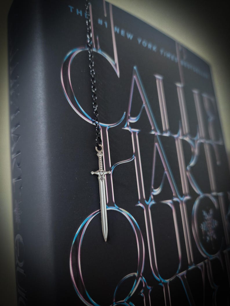 May include: A silver sword charm hangs from a black chain, resting against a dark book cover. The book's title is in large, stylized letters with a blue and pink gradient. The text at the top reads "The #1 New York Times Bestseller."