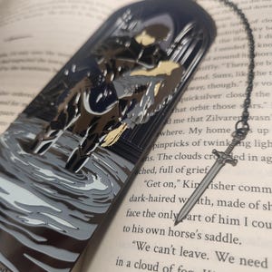 May include: A metal bookmark with a dark blue arched top featuring a knight on horseback in a watery scene. A silver sword charm hangs from a chain attached to the bookmark, resting on the open pages of a book.
