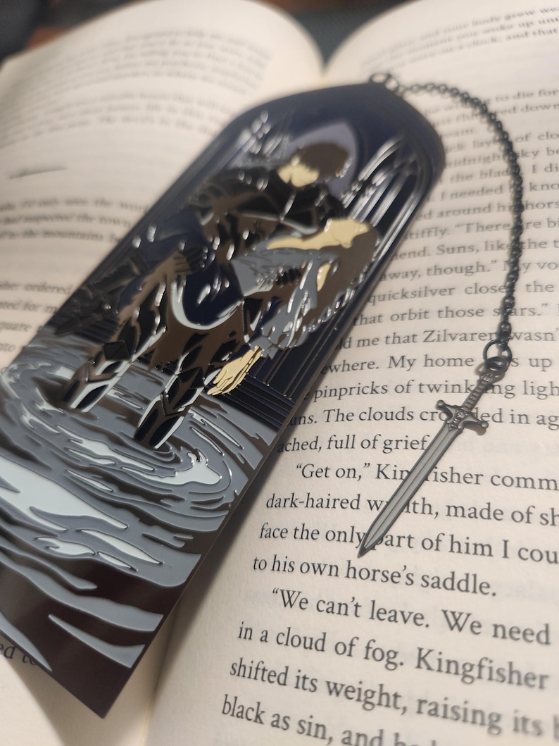 May include: A metal bookmark with a dark blue arched top featuring a knight on horseback in a watery scene. A silver sword charm hangs from a chain attached to the bookmark, resting on the open pages of a book.