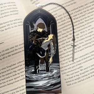 May include: Black metal bookmark with a gothic arch design. The bookmark features a detailed illustration of a man carrying a woman in water. A sword-shaped charm hangs from a chain.