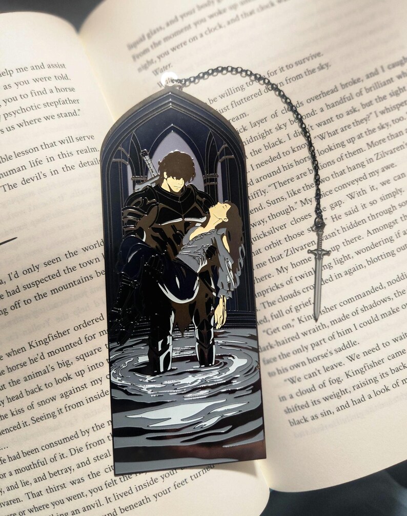 May include: Black metal bookmark with a gothic arch design. The bookmark features a detailed illustration of a man carrying a woman in water. A sword-shaped charm hangs from a chain.