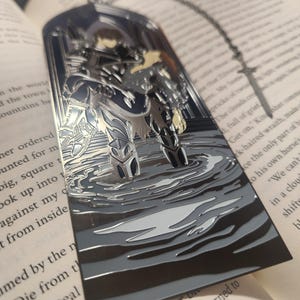 May include: A silver metal bookmark featuring a detailed illustration of a figure in armor standing in water. The bookmark has a chain attached and is set against the backdrop of an open book with printed text.