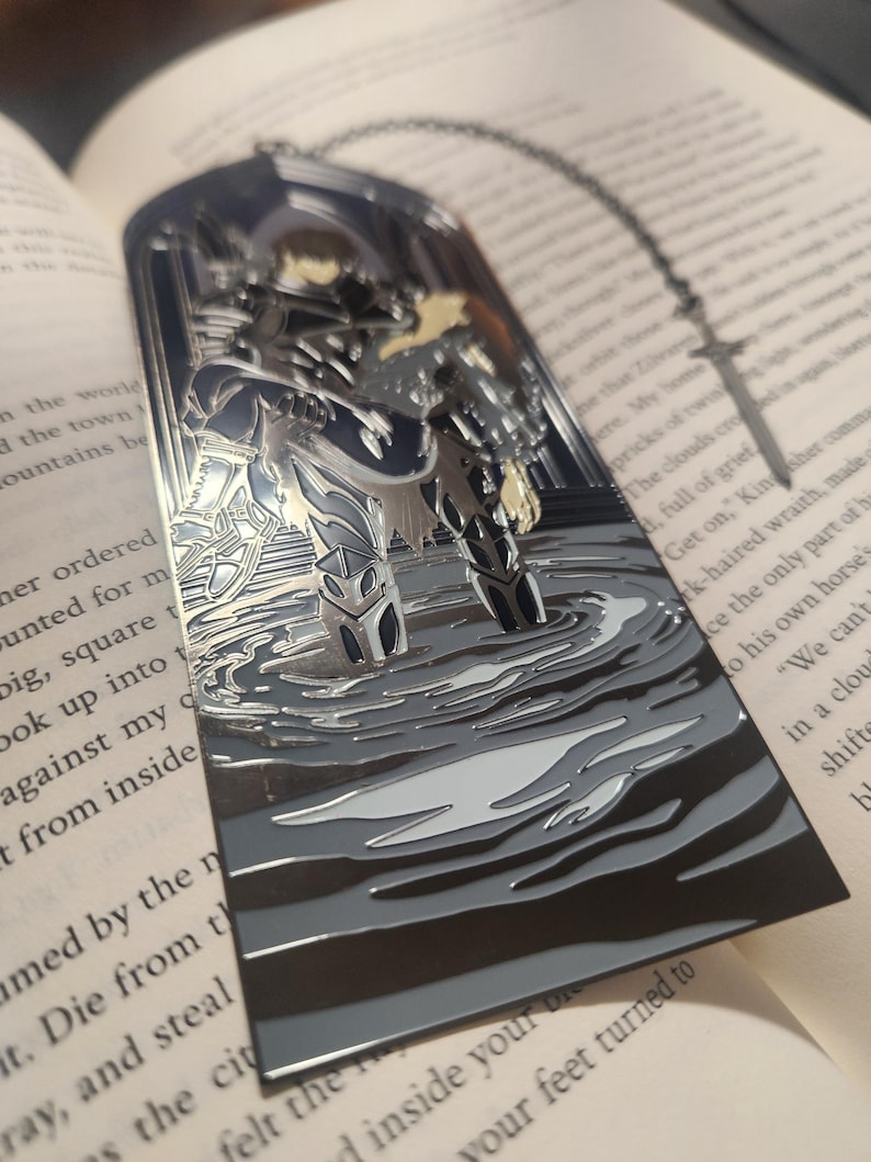 May include: A silver metal bookmark featuring a detailed illustration of a figure in armor standing in water. The bookmark has a chain attached and is set against the backdrop of an open book with printed text.