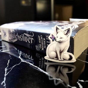 K&ouml;nnte beinhalten: Ein wei&szlig;er Fuchssticker mit schwarzspitzem Schwanz und Ohren, der einen lila Schmetterling auf der Nase h&auml;lt, sitzt auf einem Buch mit dem Titel "Quicksilver". Das Buch liegt auf einer schwarz-wei&szlig; marmorierten Oberfl&auml;che.
