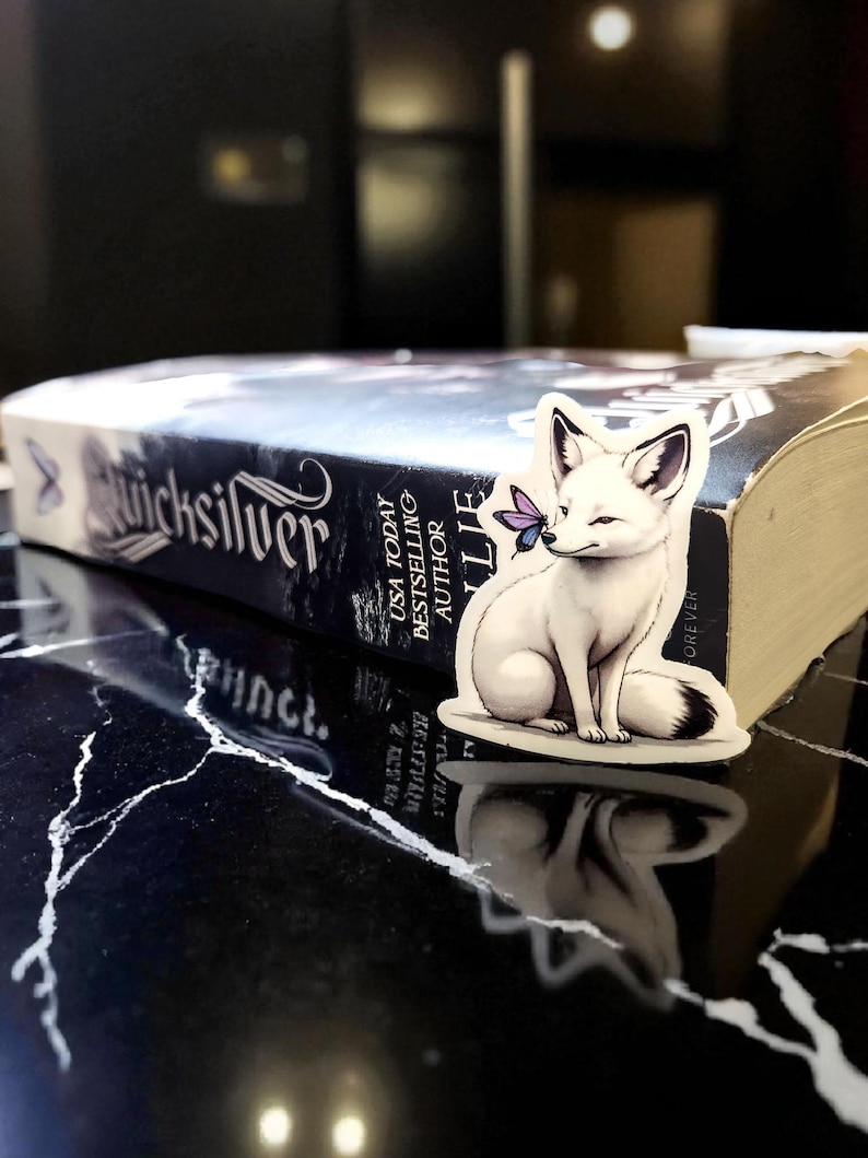 K&ouml;nnte beinhalten: Ein wei&szlig;er Fuchssticker mit schwarzspitzem Schwanz und Ohren, der einen lila Schmetterling auf der Nase h&auml;lt, sitzt auf einem Buch mit dem Titel "Quicksilver". Das Buch liegt auf einer schwarz-wei&szlig; marmorierten Oberfl&auml;che.