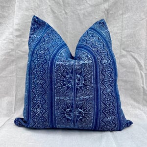 Puede incluir: Un cojín cuadrado con un patrón geométrico azul y blanco. El cojín presenta un diseño repetitivo de formas y líneas estilizadas, creando una apariencia simétrica y decorativa. El cojín está hecho de tela.