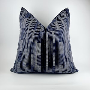 May include: A square decorative pillow with a navy blue and white geometric pattern. The pillow features a textured design with vertical and rectangular shapes, creating a modern aesthetic. The pillow is placed against a white background.