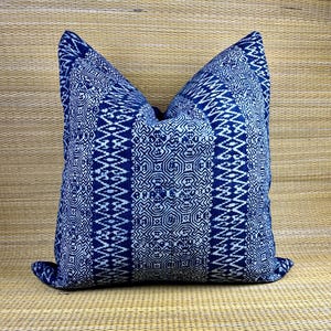 May include: Square throw pillow with a geometric pattern in shades of indigo and white. The pillow features a repeating design of lines and shapes, creating a textured appearance. The pillow is placed on a woven surface.