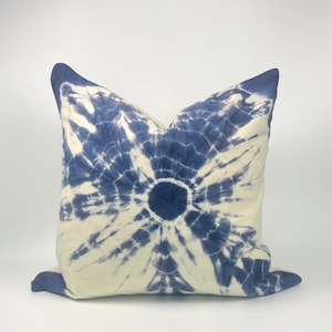 Może przedstawiać: Kwadratowa poduszka z wzorem tie-dye w odcieniach niebieskiego i kremowego. Wzór promieniuje z centralnego, ciemnoniebieskiego okręgu, tworząc efekt słońca. Poduszka ma jednolity niebieski brzeg.