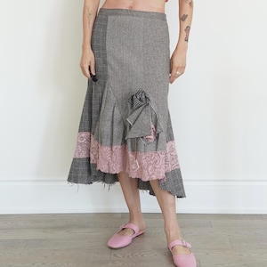 Op de afbeelding: Een grijze en zwarte tweed rok met een patchwork ontwerp. De rok heeft een gedeelte van zwart-wit ruit, een roze kanten rand en een decoratieve stoffen bloem. De zoom is ongelijkmatig, wat het een unieke, vintage look geeft.