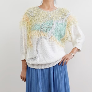 May include: A white knitted sweater with a colourful, beaded design on the front. The design features shades of yellow, green, and silver, with a variety of beads and sequins. The sweater has a fluffy texture and a relaxed fit.