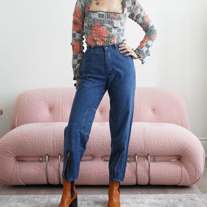 Könnte beinhalten: Eine Person trägt ein gemustertes Langarm-Top mit quadratischem Ausschnitt und eine hoch taillierte blaue Jeans. Die Jeans hat Kniepatches und Reißverschlüsse an den Knöcheln. Sie trägt braune Leder-Ankle Boots mit schwarzen Absätzen.