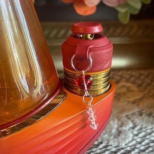 May include: A red and gold vaporizer with a heart-shaped charm. The vaporizer has a gradient colour scheme, transitioning from red to orange. A small, red heart charm and clear crystal beads dangle from the device.