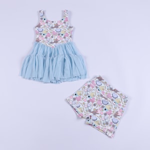 Può includere: Un vestito azzurro con gonna in tulle e pantaloncini abbinati. L'abito e i pantaloncini presentano un motivo di immagini di cartoni animati rosa, gialli e blu su sfondo bianco. L'abito ha un top senza maniche.