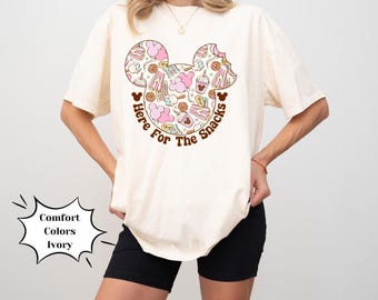May include: A white t-shirt with a pink and brown Mickey Mouse head design. The Mickey Mouse head is filled with images of snacks and the text "Here For The Snacks".