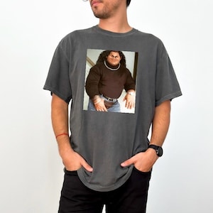 Op de afbeelding: Een donkergrijs T-shirt met een bedrukte afbeelding van een man die een bruine trui en jeans draagt. De man heeft lang zwart haar en een dikke zwarte snor.