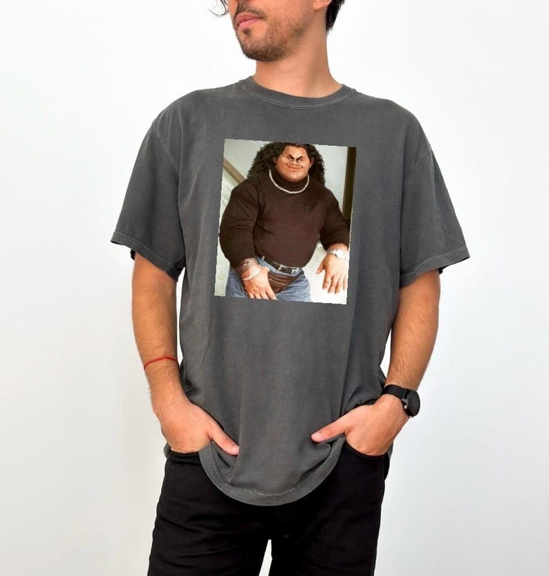 Op de afbeelding: Een donkergrijs T-shirt met een bedrukte afbeelding van een man die een bruine trui en jeans draagt. De man heeft lang zwart haar en een dikke zwarte snor.