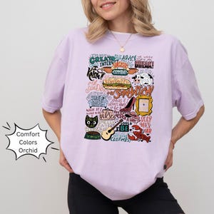 Op de afbeelding: Een lichtpaarse T-shirt met een grafische print van verschillende zinnen en afbeeldingen uit de populaire tv-serie "Friends". De print bevat zinnen zoals "Ik ben niet goed in advies geven", "We waren uit elkaar", "Stinkende kat", "Kan ik nog meer zijn", en "Hoe gaat het?" De print bevat ook afbeeldingen van een kat, een kreeft, een sandwich en een gitaar.