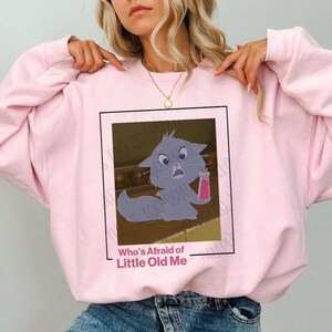 May include: Pink sweatshirt with a cartoon cat holding a pink bottle. The text "Who's Afraid of Little Old Me" is printed below the image.