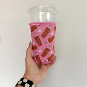 May include: A clear plastic iced coffee cup featuring a pink neoprene sleeve. The sleeve is decorated with a repeating pattern of brown iced coffee drinks with teal straws, white castle silhouettes, and small colourful stars against a pink background.