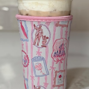 Könnte beinhalten: Eine wiederverwendbare Kaffeebecherhülle mit einem rosa-weiß gestreiften Muster. Die Hülle zeigt gerahmte Illustrationen eines Schlosses, eines Kleides und von Figuren. Der obere Rand ist mit rosa Stoff verziert. Der Becher enthält ein Getränk mit Schaum.