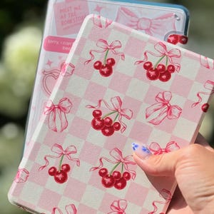 May include: A pink and white checkered case with a cherry and bow pattern. The case is held in front of a clear case with text that reads "Meet me at the bookstore" and "Sorry, I can't be there."